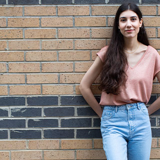 A student leaning against a brick wall, smiling at the camera. 